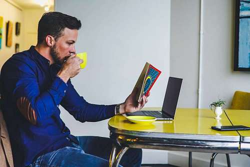 young man has breakfast while reading a book in front of his laptop